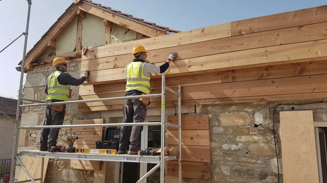 Des artisans sont en train de poser des lames de bardage en bois sur l'échafaudage d'une maison en rénovation, mettant en avant l'aspect esthétique et la durabilité de ce matériau. La façade du bâtiment prend forme, offrant une solution de revêtement résistante aux intempéries.