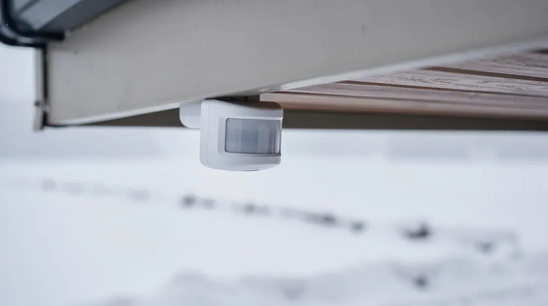 Un détecteur de mouvement extérieur est fixé sous un avant-toit, tandis que la neige recouvre le sol en contrebas, créant un contraste entre l'équipement technologique et l'environnement hivernal. Cet appareil est conçu pour détecter la présence de personnes et assurer une efficacité optimale dans diverses applications.