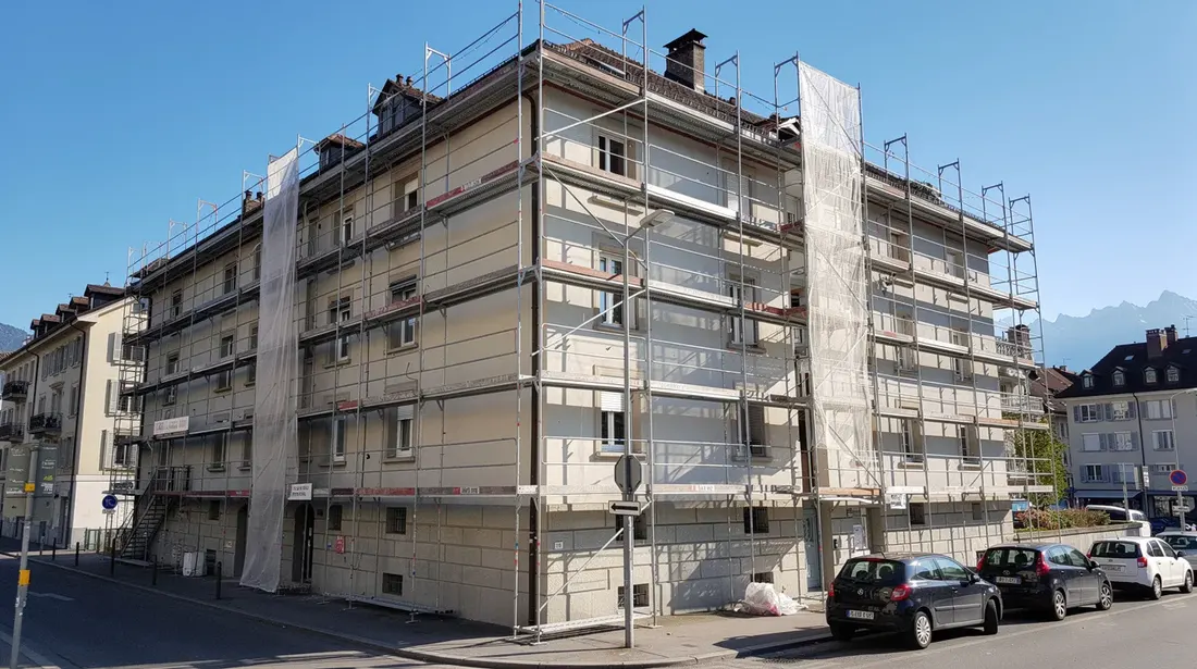L'image montre un bâtiment résidentiel genevois en cours de rénovation, entouré d'échafaudages, sous un ciel bleu éclatant. Ce projet s'inscrit dans le cadre des efforts de rénovation du canton de Genève, visant à améliorer l'efficacité énergétique des bâtiments grâce à des subventions et des mesures incitatives.